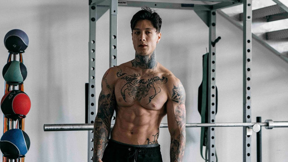 Chris Heria shirtless in a gym in front of a training rack, displaying his muscular tattooed torso and athletic build