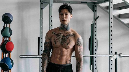 Chris Heria shirtless in a gym in front of a training rack, displaying his muscular tattooed torso and athletic build