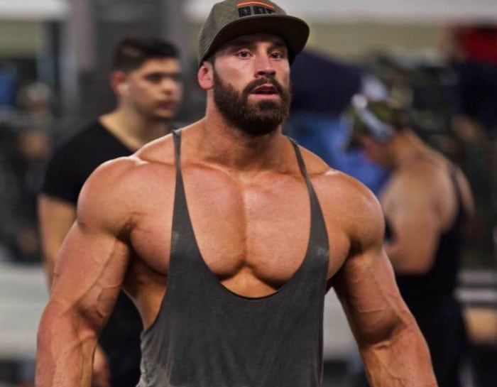 Bradley Martyn in a gym wearing a dark tank top and cap, muscular upper body visible