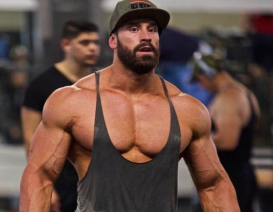 Bradley Martyn in a gym wearing a dark tank top and cap, muscular upper body visible