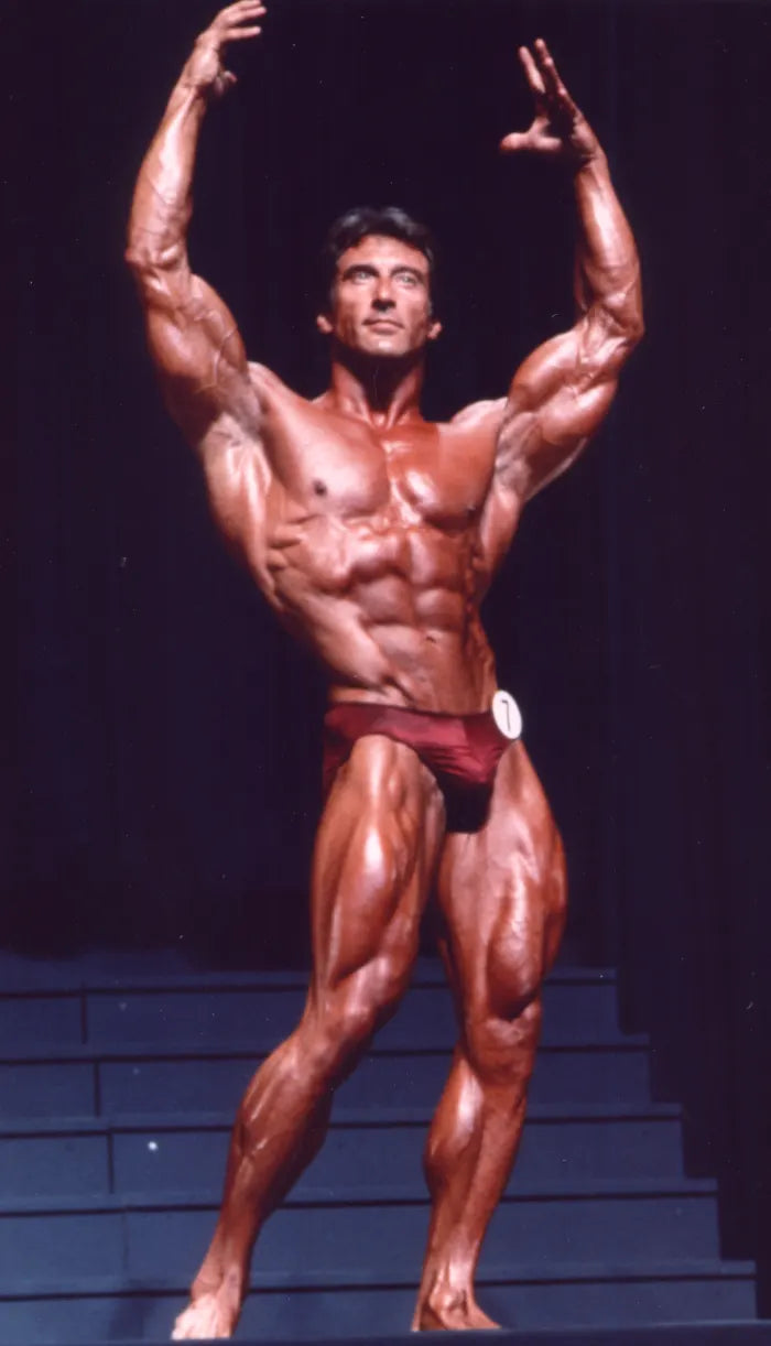 Frank Zane posing on the Mr. Olympia stage in red trunks with both arms raised