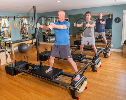 Three adult men practicing controlled movements on Pilates reformer machines in a bright studio