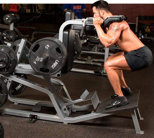 Shirtless lifter facing the pads of a reverse hack squat machine, squatting with hips back and feet planted