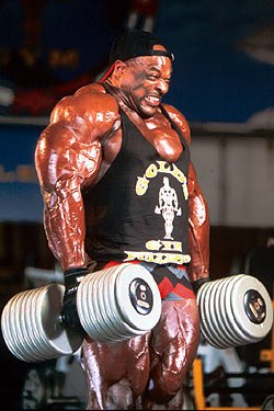 Prime-era Ronnie Coleman performing a heavy dumbbell shrug, showing massive trapezius and shoulder development