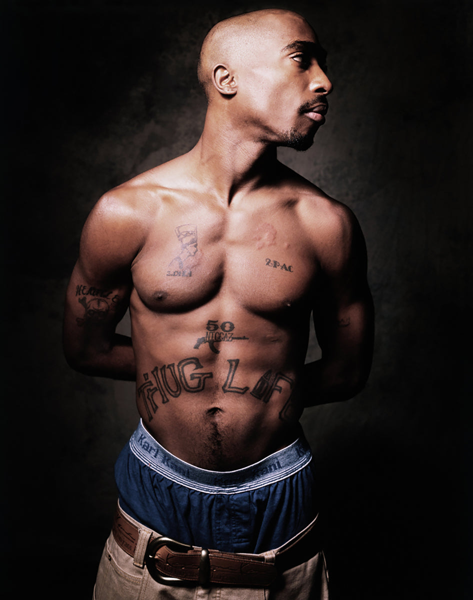 Color studio portrait of Tupac Shakur shirtless with defined shoulders, chest, abdomen and THUG LIFE tattoo