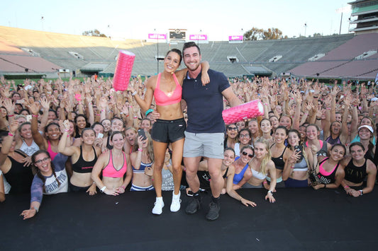 Kayla Itsines and a fellow trainer holding pink foam rollers before a cheering outdoor fitness crowd
