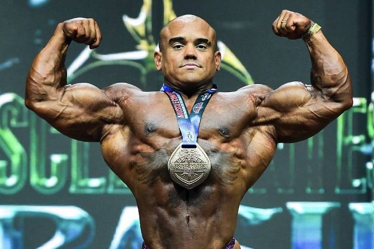 Vitor Porto flexes in a front double-biceps pose onstage with a winner's medal after his 2026 Olympia Brasil 212 victory