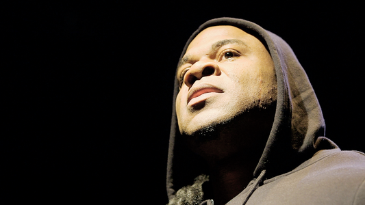 Close-up of Kai Greene in a dark hoodie looking upward thoughtfully against a black background