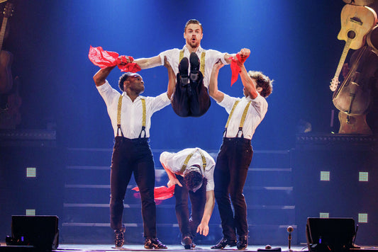 Derek Hough suspended midair in a stage dance routine with fellow performers under blue lighting
