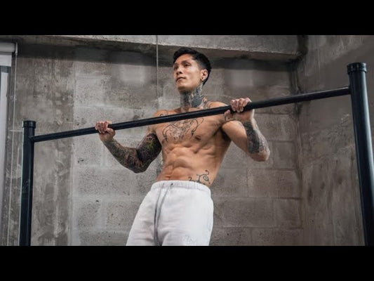Chris Heria performing a strict pull-up on a horizontal bar in a concrete-walled training space