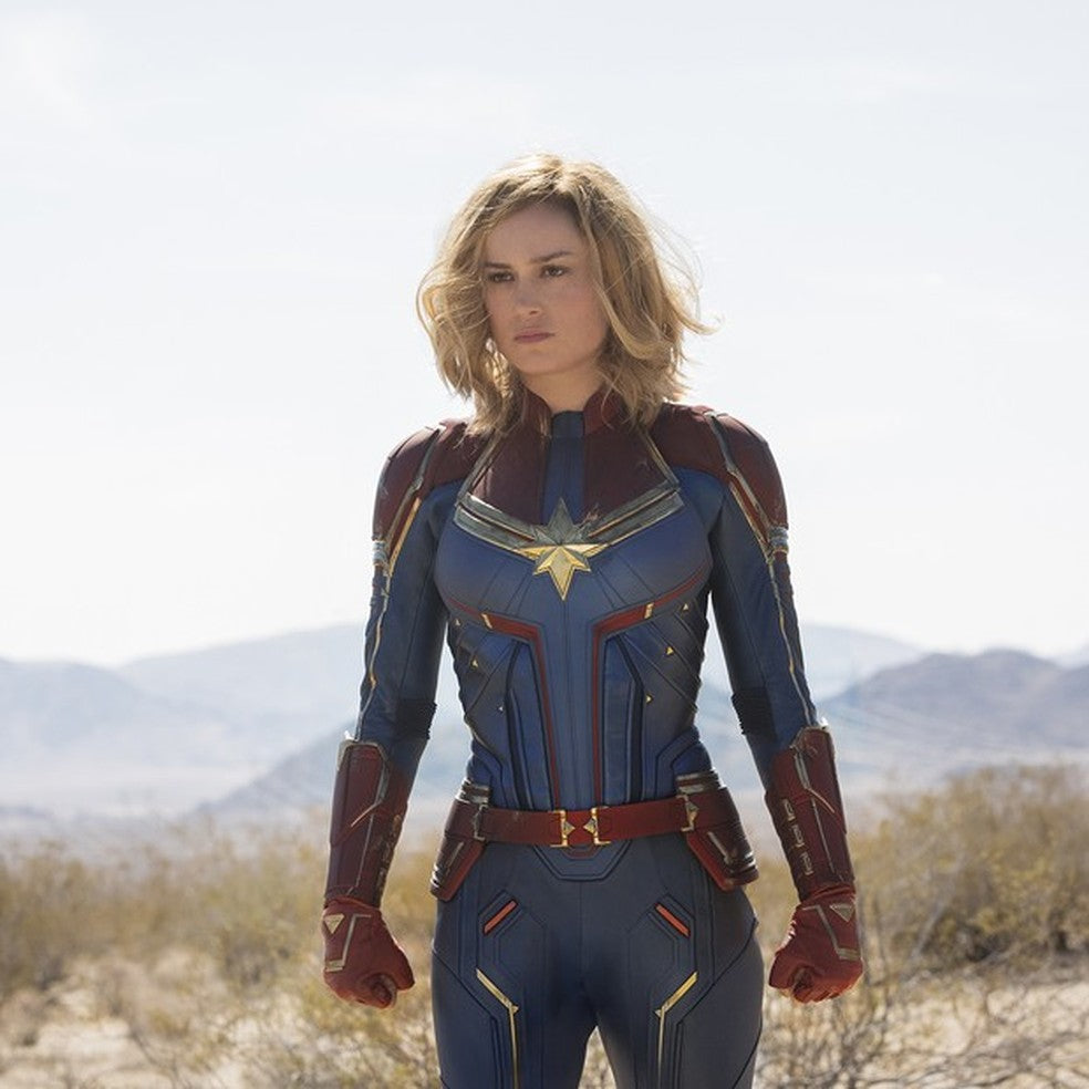 Brie Larson as Captain Marvel in a blue, red and gold superhero suit standing in a desert