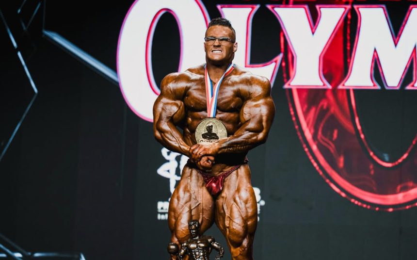Bodybuilder holding an Olympia medal and trophy onstage after earning the sport’s biggest title