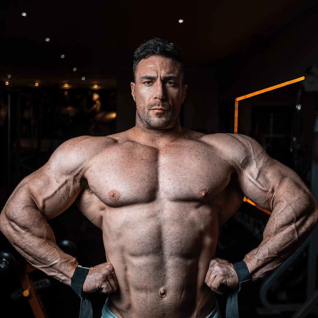 Noel Deyzel shirtless in a gym with arms braced on hips and broad rounded deltoids clearly visible