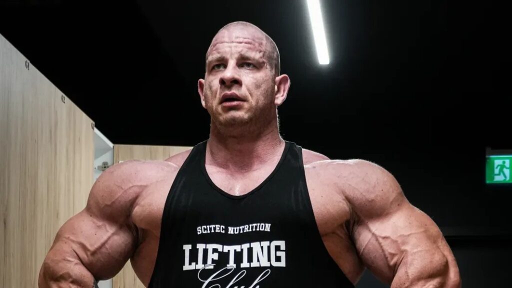 Michal Krizo wearing a black training tank top in a gym, displaying massive shoulders and arms