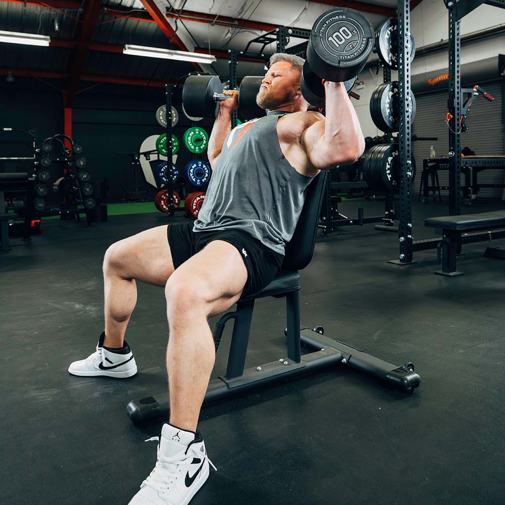 Real athlete seated on a bench pressing dumbbells in a functional gym, visible weight plates and rack; no text overlay