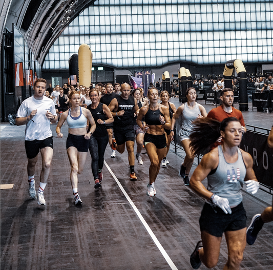 Competitors launch from the start of an indoor HYROX fitness race under an illuminated event arch.