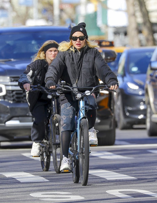 Madonna cycling on a blue bicycle in a black jacket and beanie on a city street