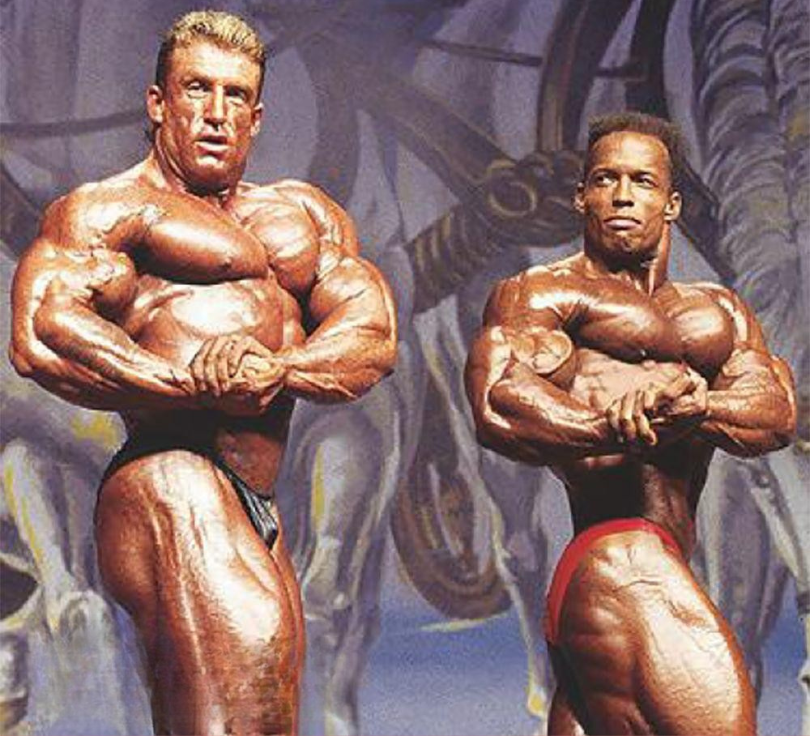 Dorian Yates and Shawn Ray posing side by side in the side chest at the 1994 Mr. Olympia, contrasting massive muscle thickness with compact symmetry
