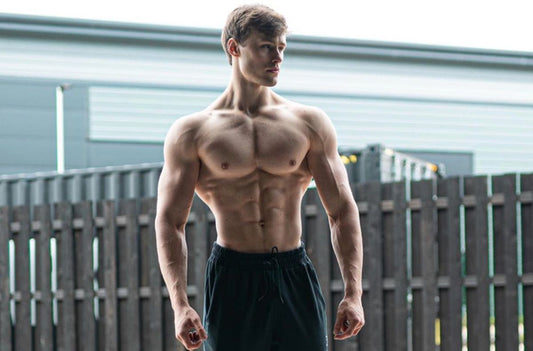 David Laid standing shirtless with broad shoulders, developed chest and visible narrow waist in front of a wooden fence