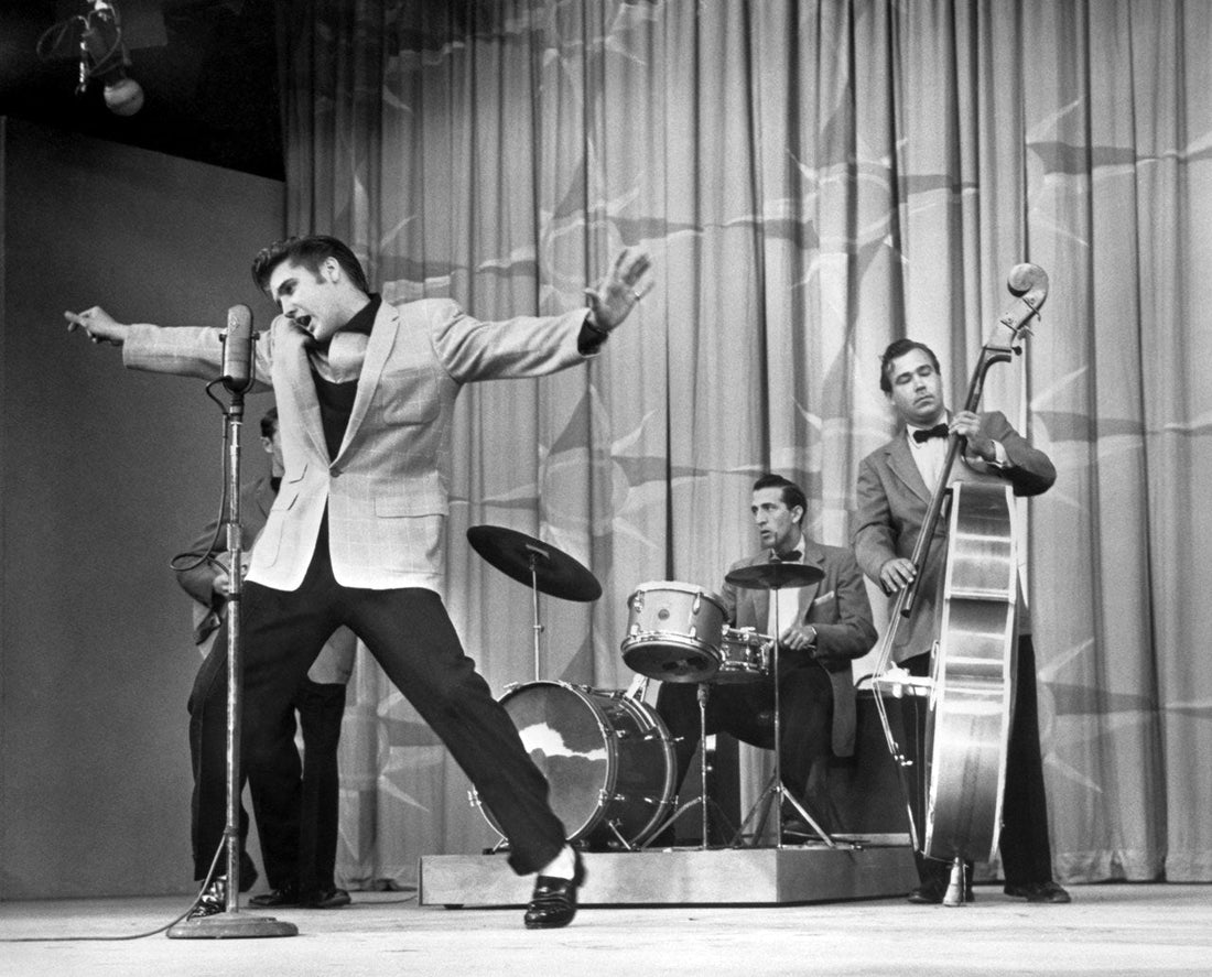 Elvis Presley performing on The Milton Berle Show in 1956, wearing a light jacket and dark trousers with his arms spread in a dynamic singing pose