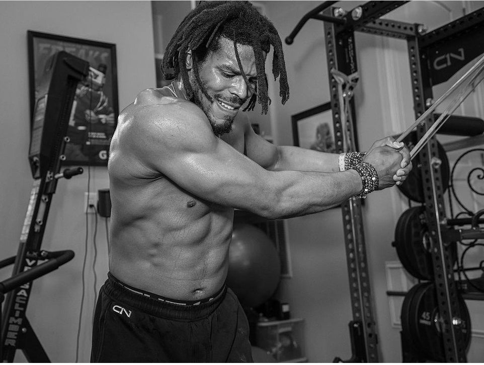 Cam Newton shirtless in a home gym performing a resistance-band core exercise
