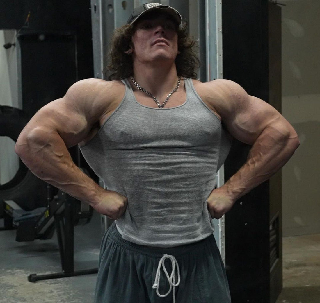 Sam Sulek standing in a gym with hands on hips, wearing a gray tank top and showing muscular arms and shoulders