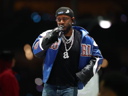 Kendrick Lamar performing live in a blue-and-black varsity jacket, holding a microphone