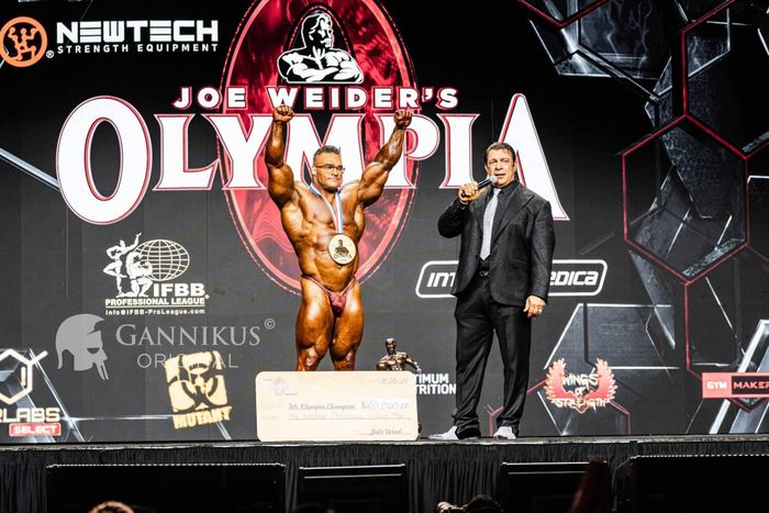 Nick Walker celebrating his 2026 Mr. Olympia victory onstage with raised arms, gold medal and trophy
