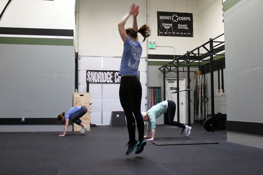 Three athletes performing burpees in a functional fitness gym, one jumping as others move through floor repetitions.