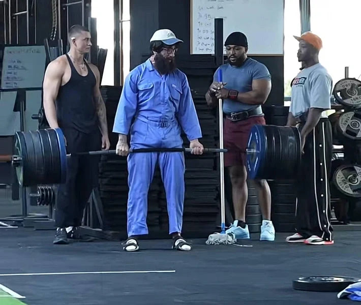 Vladimir Shmondenko dressed as a blue-uniformed gym cleaner with a heavy barbell and muscular gymgoers nearby