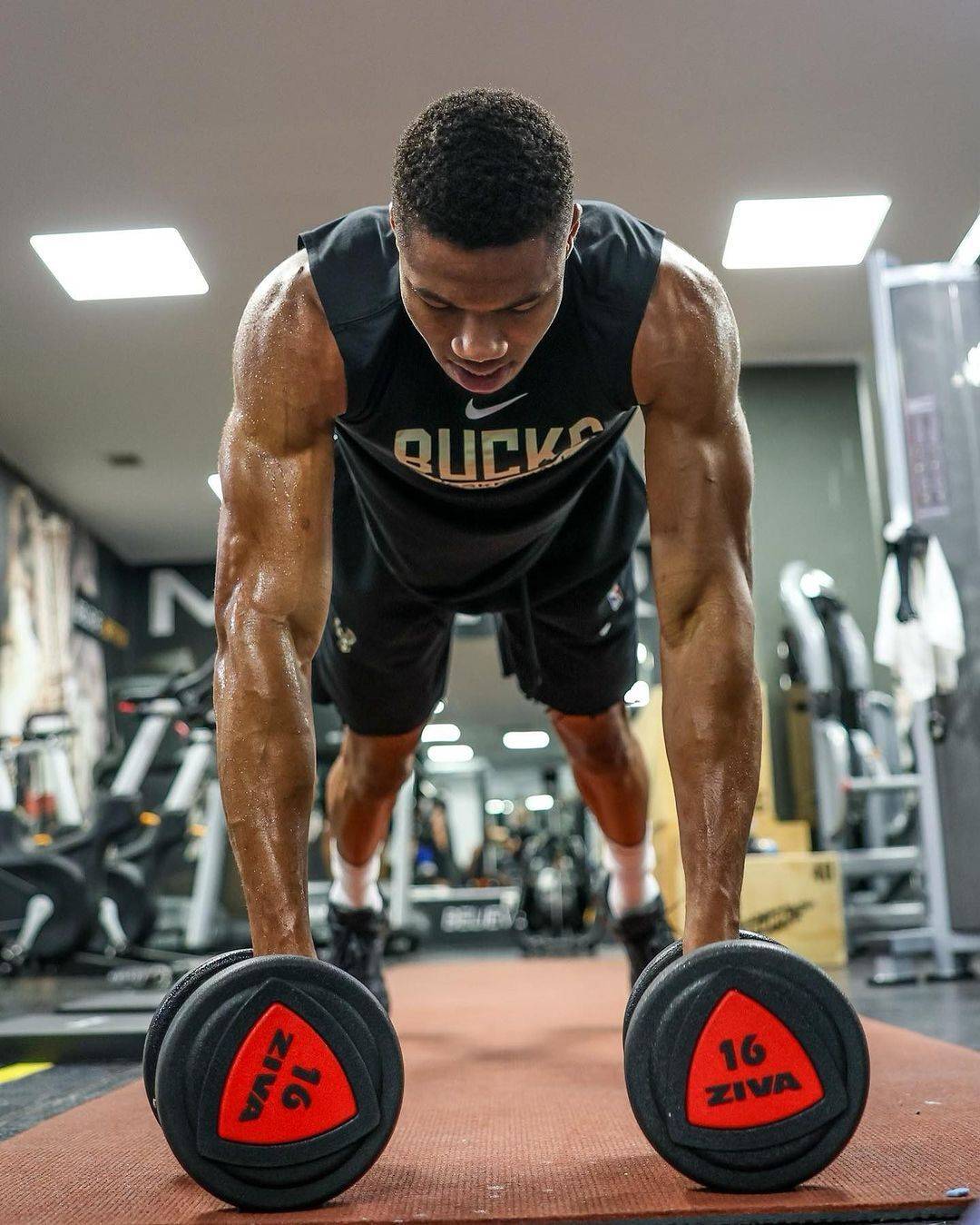 Giannis Antetokounmpo performing a demanding push-up on two dumbbells in a Milwaukee Bucks training gym