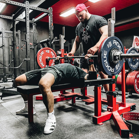 Powerlifter bench pressing a heavily loaded barbell with a spotter in a training gym