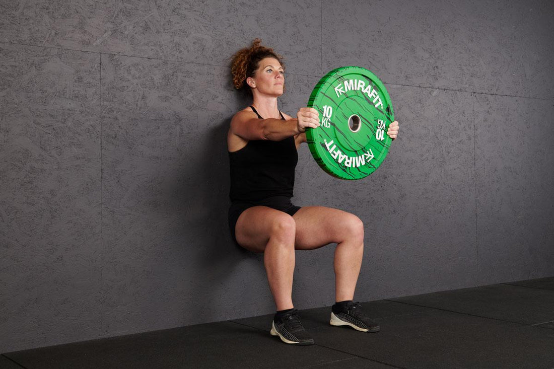 Woman in black gym clothing performing a weighted wall sit while holding a green 10 kg plate against a grey wall