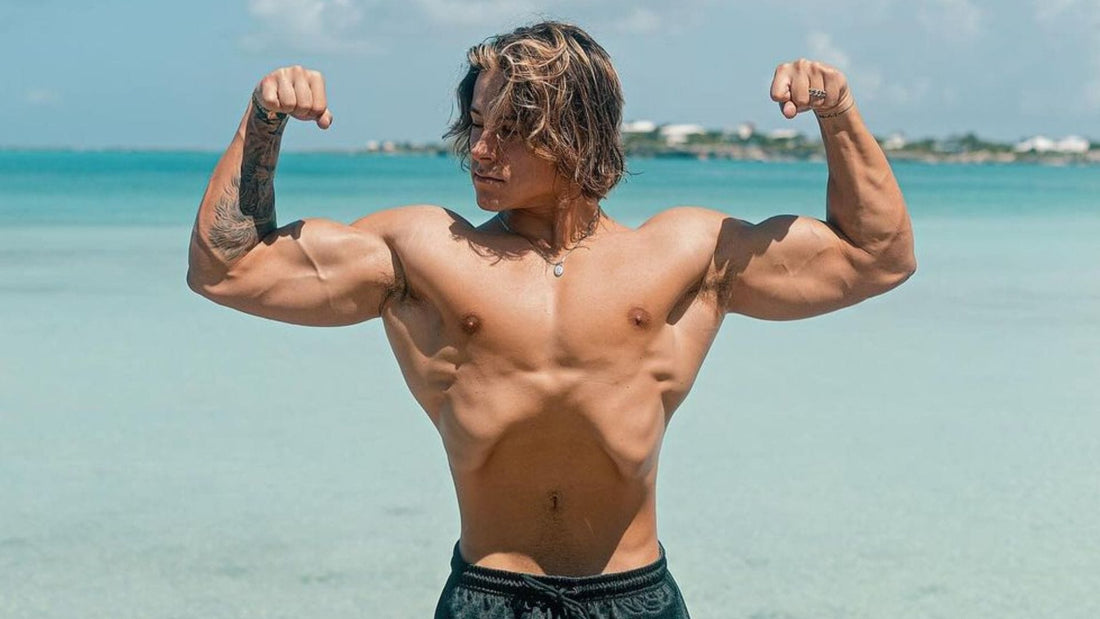 Alex Eubank flexing his arms and chest on a beach, showing a lean aesthetic bodybuilding physique
