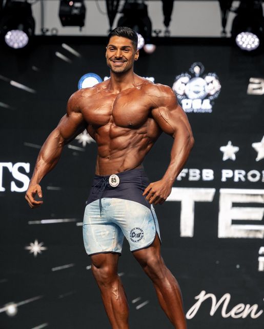 Andrei Deiu smiling on the 2026 Olympia Men’s Physique stage with visibly defined shoulders, chest and abdominals