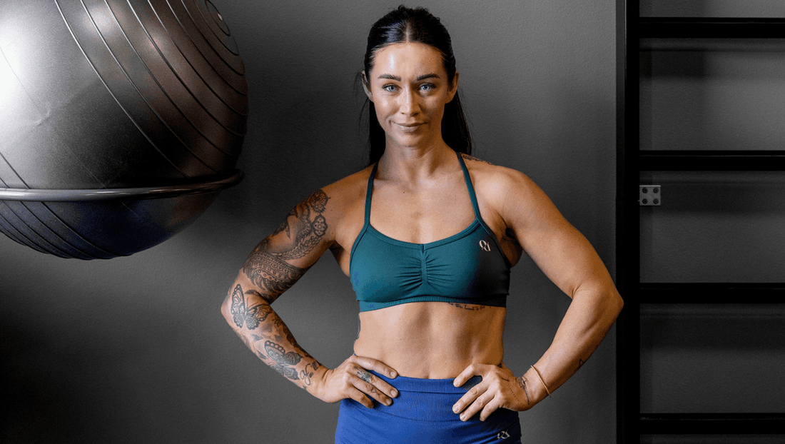 Hanna Öberg standing in teal sports bra and blue leggings, hands on hips, showing muscular arms and core