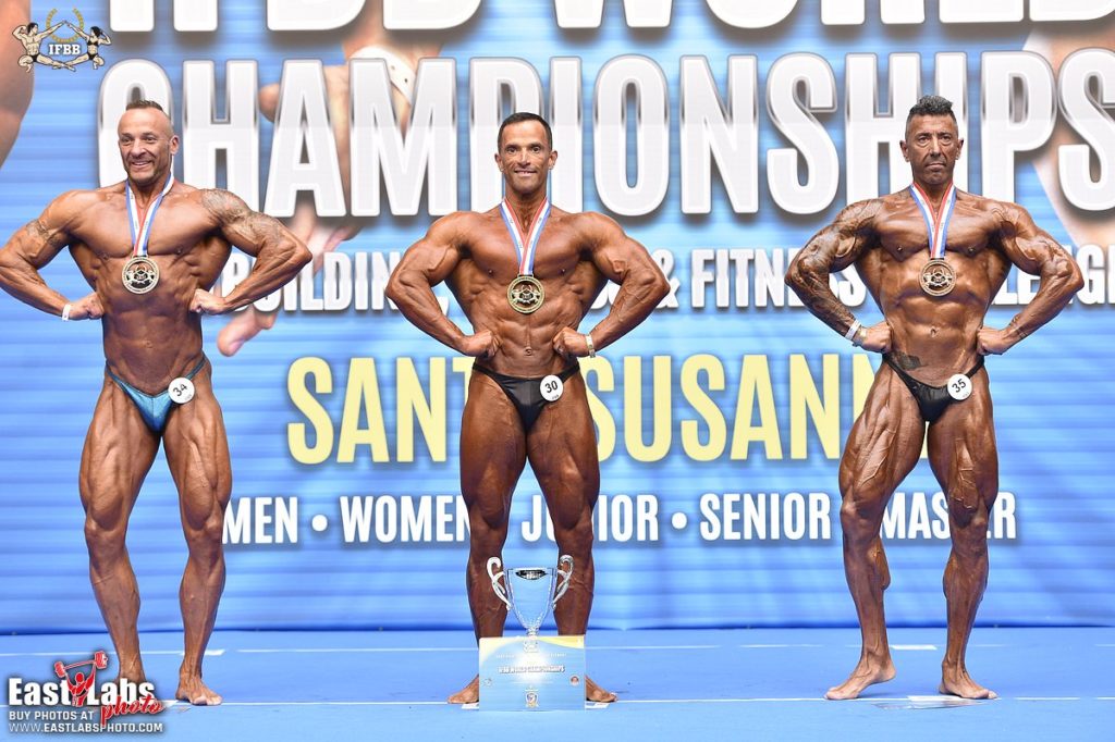 Three male bodybuilding competitors on a blue IFBB championship stage, the center athlete standing behind a trophy