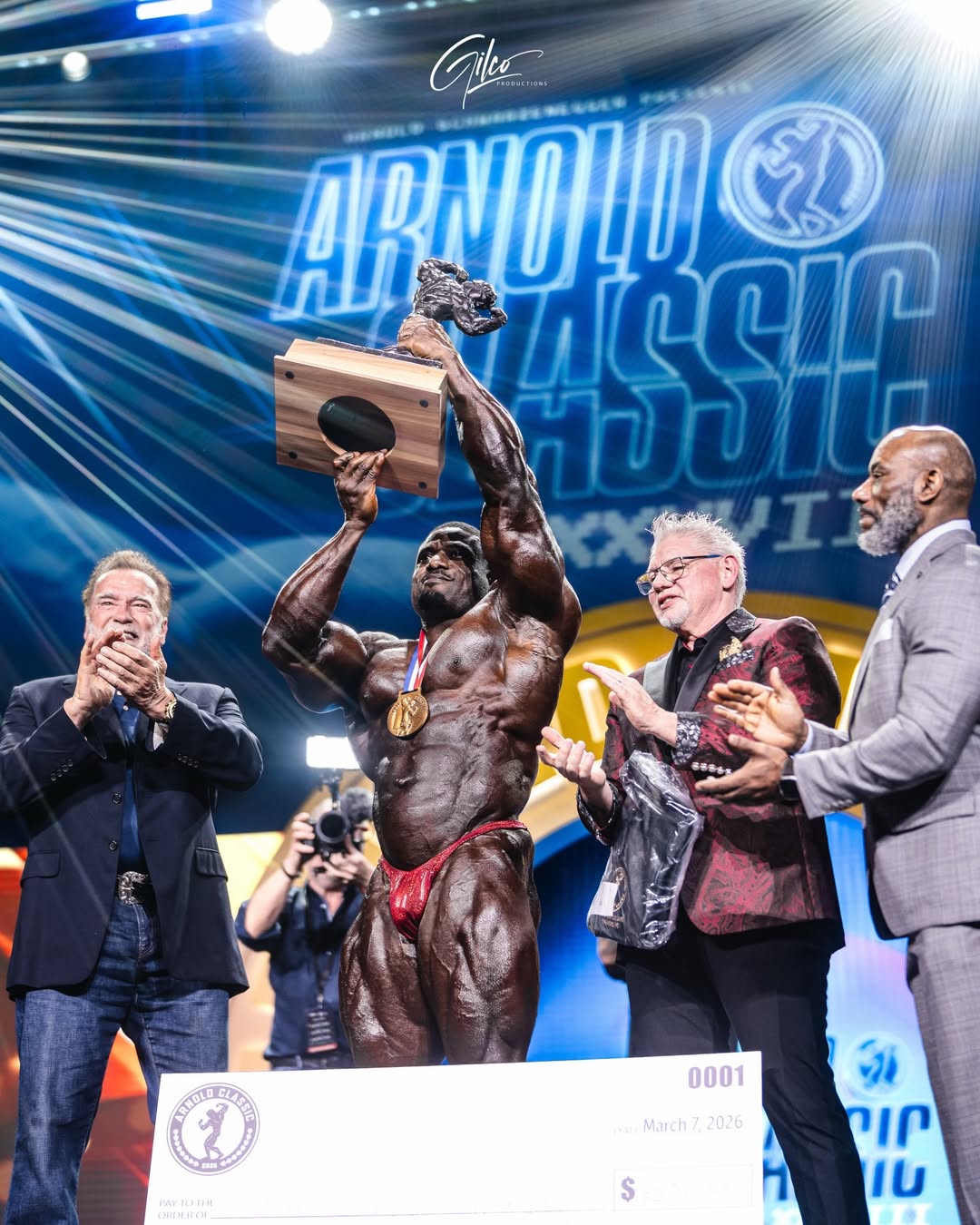 Andrew Jacked lifting the 2026 Arnold Classic trophy onstage as Arnold Schwarzenegger applauds