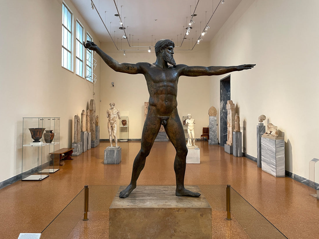 Ancient bronze statue of a muscular bearded Greek god with outstretched arms inside an Athens museum; traditionally identified as Zeus or Poseidon