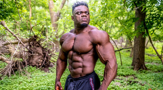 Blessing Awodibu posing shirtless with defined chest and arms in a green outdoor setting