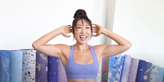 Cassey Ho smiling in lavender workout top while demonstrating a standing arms exercise in a bright home studio
