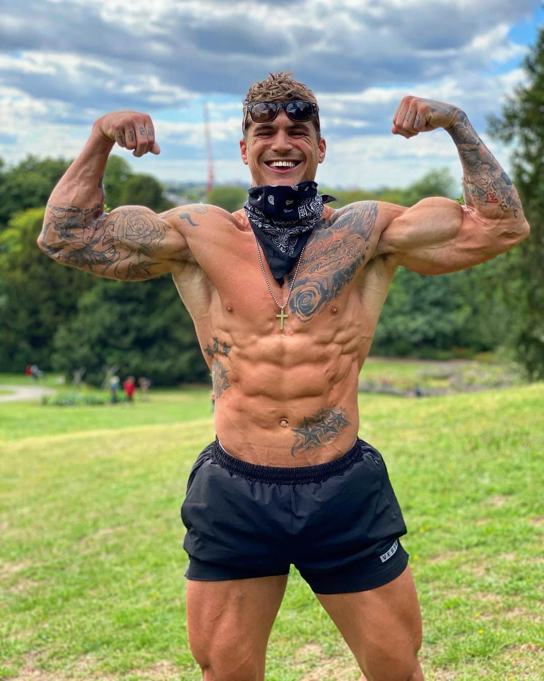 Ross Dickerson smiling while flexing both arms outdoors, showing developed biceps and lean physique