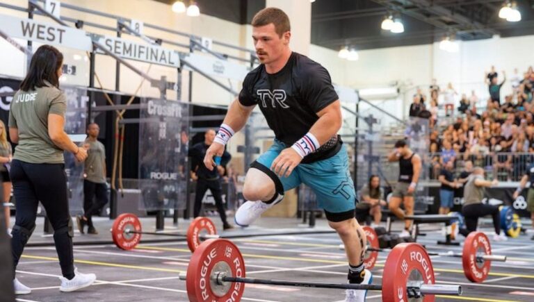 CrossFit competitor jumping over a barbell during a burpee workout in a competition gym