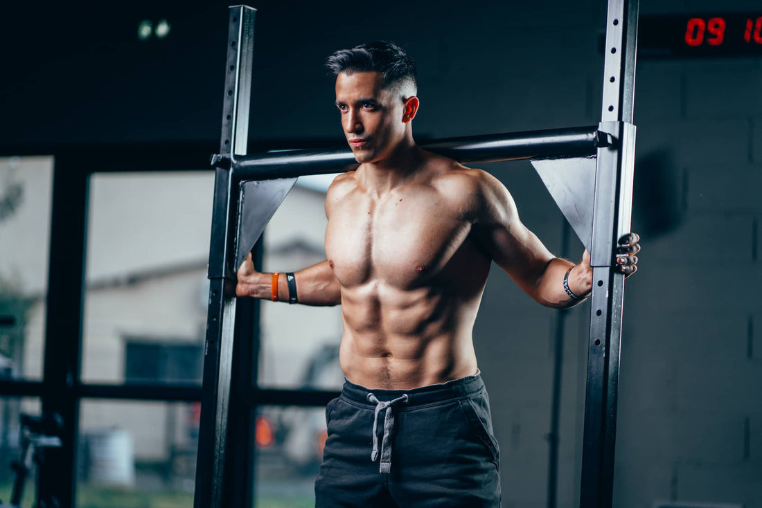 Tibo InShape standing shirtless beside a gym rack, showing developed chest and shoulders