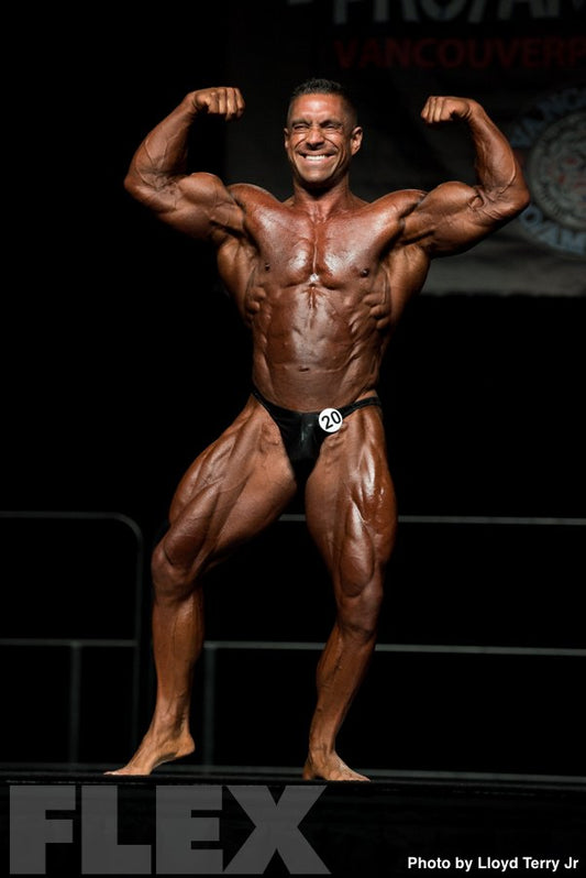Greg Doucette flexing both biceps on a professional bodybuilding stage