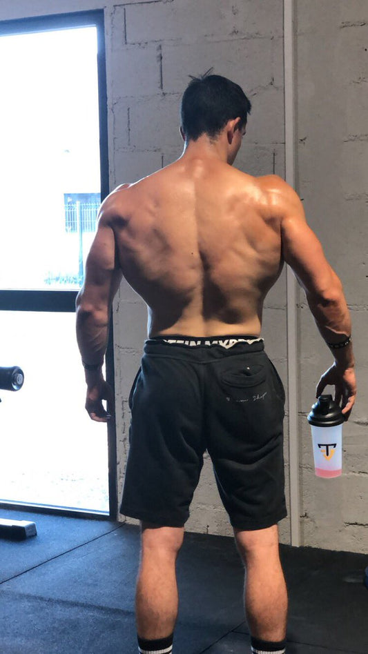 Tibo InShape shows muscular back width and a pronounced shoulder-to-waist taper while standing shirtless in a gym