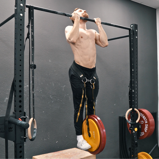 An athlete performing a heavy weighted pull-up with a weight plate hanging from a dip belt inside a gym rack