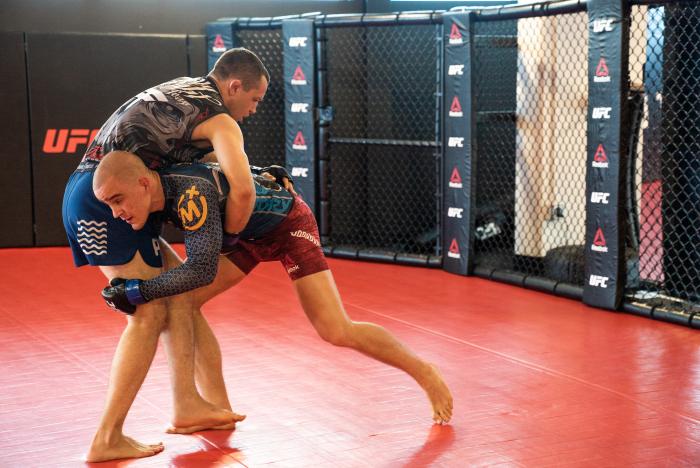 Two mixed martial arts athletes wrestling and drilling a takedown on a red mat inside a UFC training facility