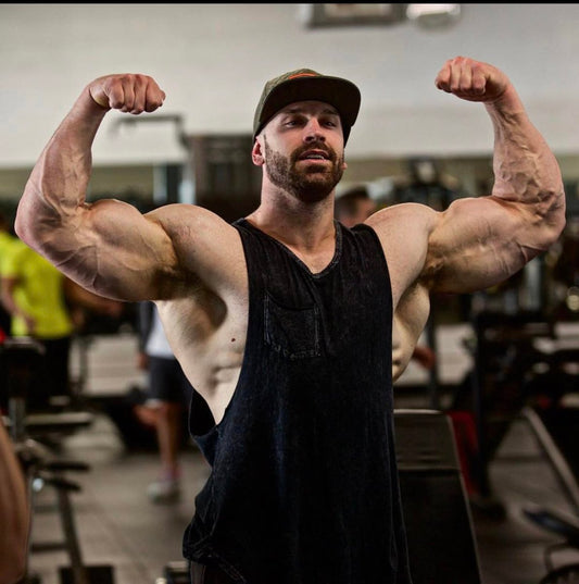 Bradley Martyn flexing both arms in a black tank top on a busy gym floor