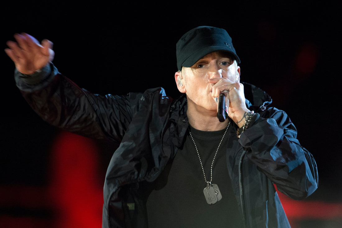 Eminem performing onstage in a gray hooded sweatshirt and cap while holding a microphone